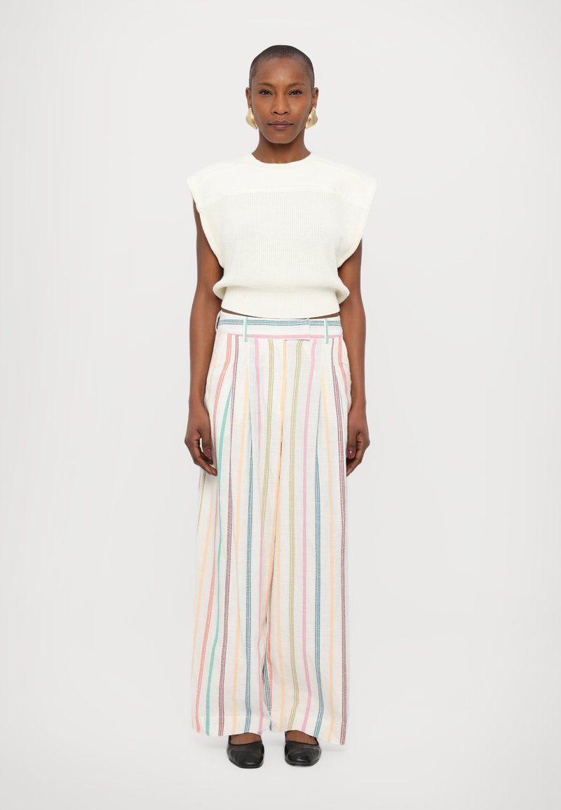 Svetlo pletena brezrokavna majica v off-white barvi, ujemajoča se s široko prugastimi hlačami pastelnih barv, ki imajo pas s pasom in sproščen kroj.