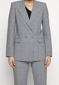 Šedý dvouřadý blazer s kostkovaným vzorem, který obsahuje růžové a zelené pruhy, přiléhavý střih, klopové límce a dvě přední kapsy.