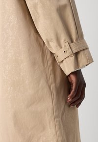Main à la peau foncée reposant à côté d'une manche de long manteau beige avec une sangle réglable et des œillets métalliques sur un fond gris.