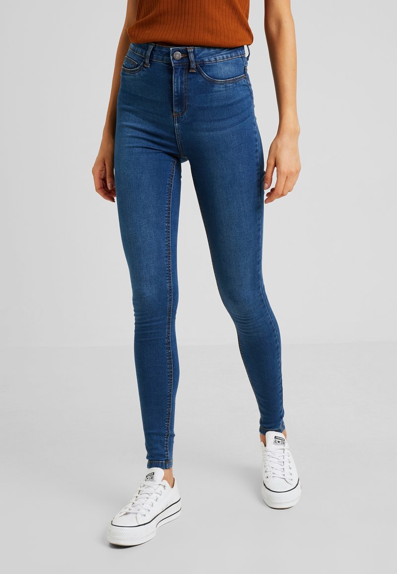 Vaqueros de mezclilla azul de cintura alta con un ajuste slim, que cuentan con un clásico diseño de cinco bolsillos y costuras en contraste, combinados con zapatillas blancas.