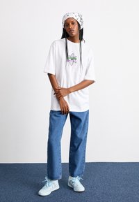 White oversized T-shirt with floral graphic, paired with light blue straight-leg jeans and pastel blue sneakers with bows. White bandana on head.