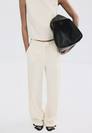 Femme portant un haut sans manches crème à fines rayures et un pantalon large assorti, tenant un grand sac en cuir noir sous le bras.