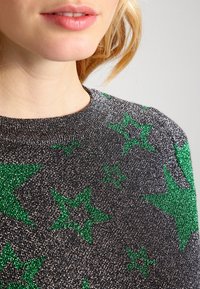 Pull noir avec un éclat métallisé arborant des motifs d'étoiles vertes, col rond et manches courtes. Tissu doux avec une finition texturée.