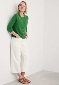 Zaļš adīts kardigans ar pogām, apvienots ar baltām platām biksēm un brūnām ādas sandalēm. Gluda tekstūra un ikdienišķs piegriezums.