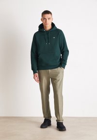 Green hoodie with drawstrings and front pocket, paired with light beige trousers and black sneakers. Simple design, cotton texture, subtle branding.