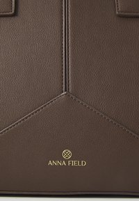 Panneau de sac en cuir marron avec surpiqûres géométriques et logo et emblème dorés "ANNA FIELD" au centre en bas.