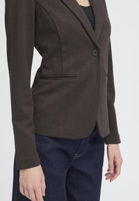 Blazer marrone a spina di pesce con chiusura a un bottone, rever a incavo e tasche laterali, abbinato a jeans denim scuro.