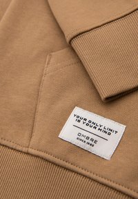 Sweat-shirt beige avec poignets côtelés et poche, avec une étiquette blanche indiquant « VOTRE SEUL LIMITEST VOTRE ESPRIT » et « OMBRE depuis 1985 ».