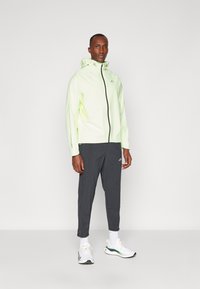Neon zelena lagana jakna s patentnim zatvaračem, kapuljačom i reflektirajućim detaljima, u kombinaciji s tamno sivim tapered hlačama i bijelim sportskim cipelama.