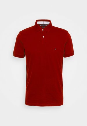 Red polo shirt with short sleeves, collar, and two-button placket. Features a small logo on the left chest, smooth fabric texture.