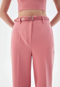 Woman wearing high-waisted pink trousers with matching belt and cropped pink top, hands resting on hips, shown from waist to mid-thigh.