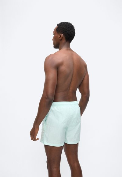 Άνδρας με σκουρόχρωμο δέρμα που φοράει ανοιχτοπράσινα μαγιό στέκεται με την πλάτη στην κάμερα σε λευκό φόντο.