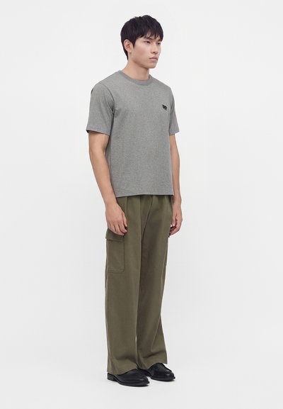 Modèle masculin debout portant un t-shirt gris à manches courtes et un pantalon cargo vert olive avec des chaussures noires sur un fond blanc uni.