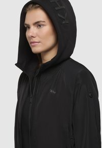 Veste légère noire avec capuche, fermeture éclair, poignets côtelés et détail de logo sur la poitrine et la manche. Texture lisse, design élégant.