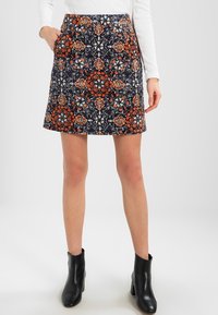 Femme portant une jupe évasée bleu marine avec un motif floral orange, blanc et beige, un haut blanc à manches longues et des bottines noires.