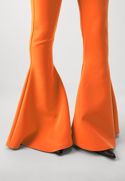 Flared trousers in bright orange fabric, featuring a smooth texture and seam detailing, paired with pointed black footwear.