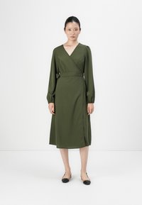 Olive green wrap dress with long sleeves, V-neckline, and a fitted waist. Smooth texture, knee-length with a side slit, paired with black flats.