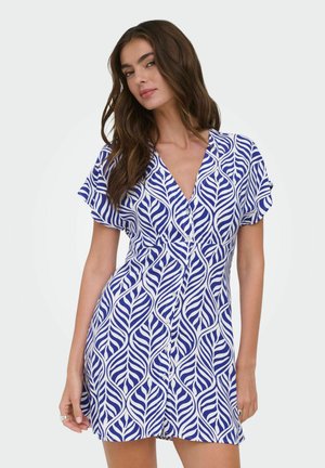 Femme portant une robe à manches courtes bleue et blanche à motifs de feuilles avec des boutons sur le devant, debout devant un fond uni.