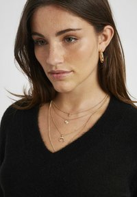 Colliers en or plaqué de longueurs et de pendentifs variés, portés sur un pull noir à col en V. Les cheveux sont ondulés doucement.