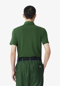 Grön pikétröja med korta ärmar, slät textur och krage. Bärs med mönstrade gröna shorts och ett mörkblått bälte med en silverspänne.