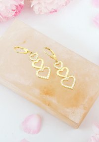 Guldhänge med tre staplade hjärtformer, två konturerade och en dekorerad med rhinestones, vilande på en ljus stenyta.