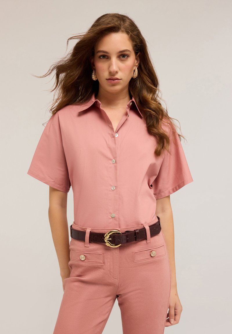 Donna con lunghi capelli ondulati indossa una camicia a maniche corte color rosa antico con bottoni, pantaloni abbinati con tasche con bottoni e una cintura scura con fibbia dorata.
