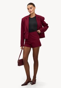 Blazer sur mesure bordeaux associé à un short taille haute assorti, top noir, escarpins à bride arrière, tenant un petit sac à main.