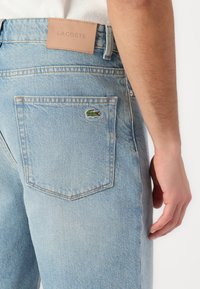 Jean en denim bleu clair avec une finition texturée, présentant une étiquette en cuir marron portant l'inscription "LACOSTE" et un logo de crocodile vert brodé sur la poche arrière.