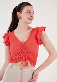 Top corto corallo con maniche corte a volant, scollo a V e arricciatura con cordoncino. Abbinato a pantaloni beige a vita alta con dettaglio a bottone.