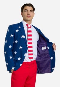 Blå blazer med hvide stjerner, åben for at afsløre lilla foer; parret med hvid skjorte, rød og hvid stribet slips og røde bukser.