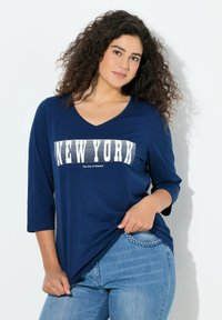 Marineblaues Langarmshirt mit V-Ausschnitt und weißem "NEW YORK"-Schriftzug, kombiniert mit hellblauen Jeans mit geflochtenem Taschen-Detail.