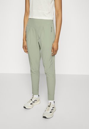 Persoon draagt lichtgroene taps toelopende broek met ritssluitingen aan de zijzakken, witte sokken, beige en zwarte sneakers en een kort wit topje.