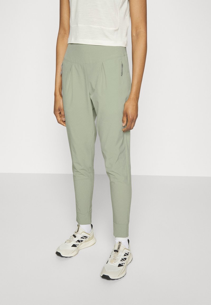 Personne portant un pantalon fuselé vert clair avec des poches latérales zippées, des chaussettes blanches, des baskets beige et noires, et un top blanc court.