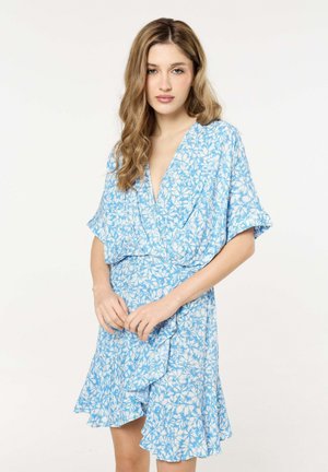 Young woman with wavy light brown hair wearing a blue and white floral wrap dress, standing with hands clasped against a plain white background.