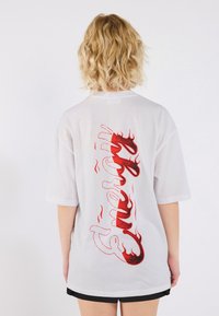 Bílý oversized tričko s krátkým rukávem a červeným grafickým motivem plamenů, na kterém je vzadu nápis "Energy". Měkká textura látky.