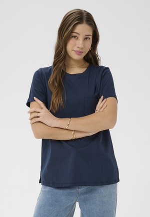 Mlada ženska z dolgimi rjavimi lasmi, ki nosi temno modro majico in svetlo modre kavbojke, z rokami prekrižanimi, z zlatim nakitom na zapestjih in prstih.