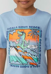 Lichtblauw t-shirt met korte mouwen featuring een afbeelding van een cartoon krokodil in een zonnebril op golven, met details van een strandscène en tekst.