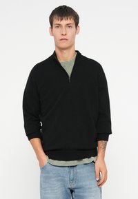 Czarny sweter zapinany na zamek z kołnierzem stójkowym i rękawami o długości trzech kwart, wykonany z miękkiego materiału, nałożony na jasnoszorty zielony shirt.