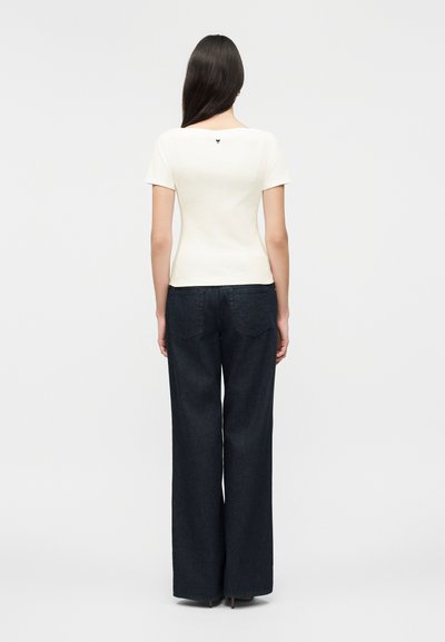 Γυναίκα με μακριά σκούρα μαλλιά φοράει στενή λευκή κοντομάνικη μπλούζα και φαρδιά σκουρόχρωμα τζιν παντελόνια, στέκεται με την πλάτη προς τον θεατή.