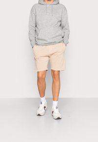 Felpa grigia con tasca frontale, abbinata a pantaloni corti color beige chiaro. Sneakers bianche con accenti rossi e blu navy. Insieme semplice e casual.
