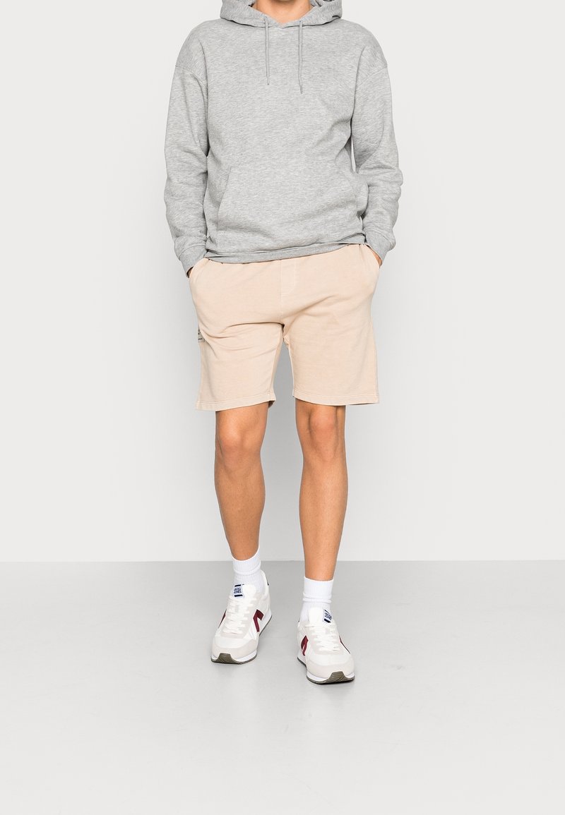 Felpa grigia con tasca frontale, abbinata a pantaloni corti color beige chiaro. Sneakers bianche con accenti rossi e blu navy. Insieme semplice e casual.