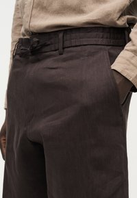 Κοντινό πλάνο ενός ατόμου που φοράει σκούρο καφέ παντελόνι με κορδόνι στη μέση και μπεζ πουκάμισο με μακριά μανίκια, με το χέρι στην τσέπη της δεξιάς πλευράς.