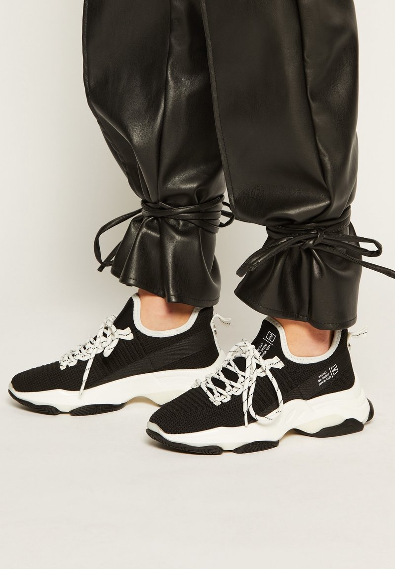 Baskets en maille noire avec semelles et lacets blancs, présentant une surface texturée et un logo sur le côté. Associées à un pantalon en similicuir noir.