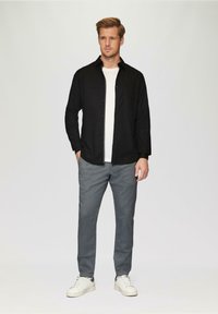 Schwarzes Hemd über einem weißen T-Shirt, kombiniert mit grauen strukturierten Hosen und weißen Turnschuhen. Einfaches und lässiges Outfit.