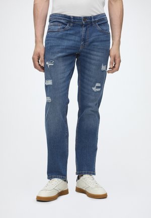 Homme portant un jean bleu usé et des baskets blanches debout devant un fond blanc uni.