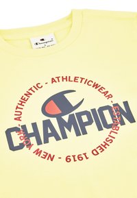 Camiseta de algodón amarillo claro con un gran logotipo negro de "CHAMPION", texto circular rojo y un pequeño logo en el cuello.