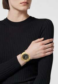 Reloj de oro con una esfera negra, correa de goma negra y acentos geométricos, llevado en una muñeca sobre un suéter negro de canalé.