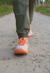 Ľahké športové topánky s bielym zvrškom a oranžovými akcentmi, textúrovanou podrážkou a zladenými šnúrkami, ktoré nosí osoba na štrkovom chodníku.