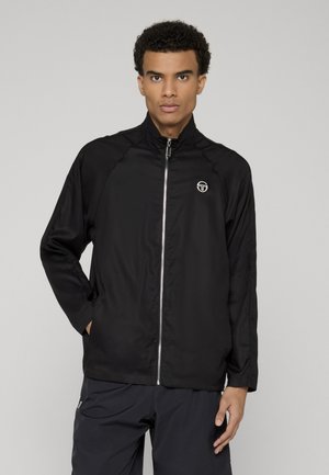 Joven con chaqueta negra con cremallera y logo en el pecho, y pantalones negros a juego, de pie contra un fondo liso de color gris claro.