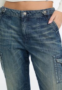 Denim-Cargo-Hosen in einer dunklen blauen Waschung, mit hohem Bund, silbernen Knöpfen, seitlichen Taschen und einer strukturierten Oberfläche mit leichten Verblassungen.